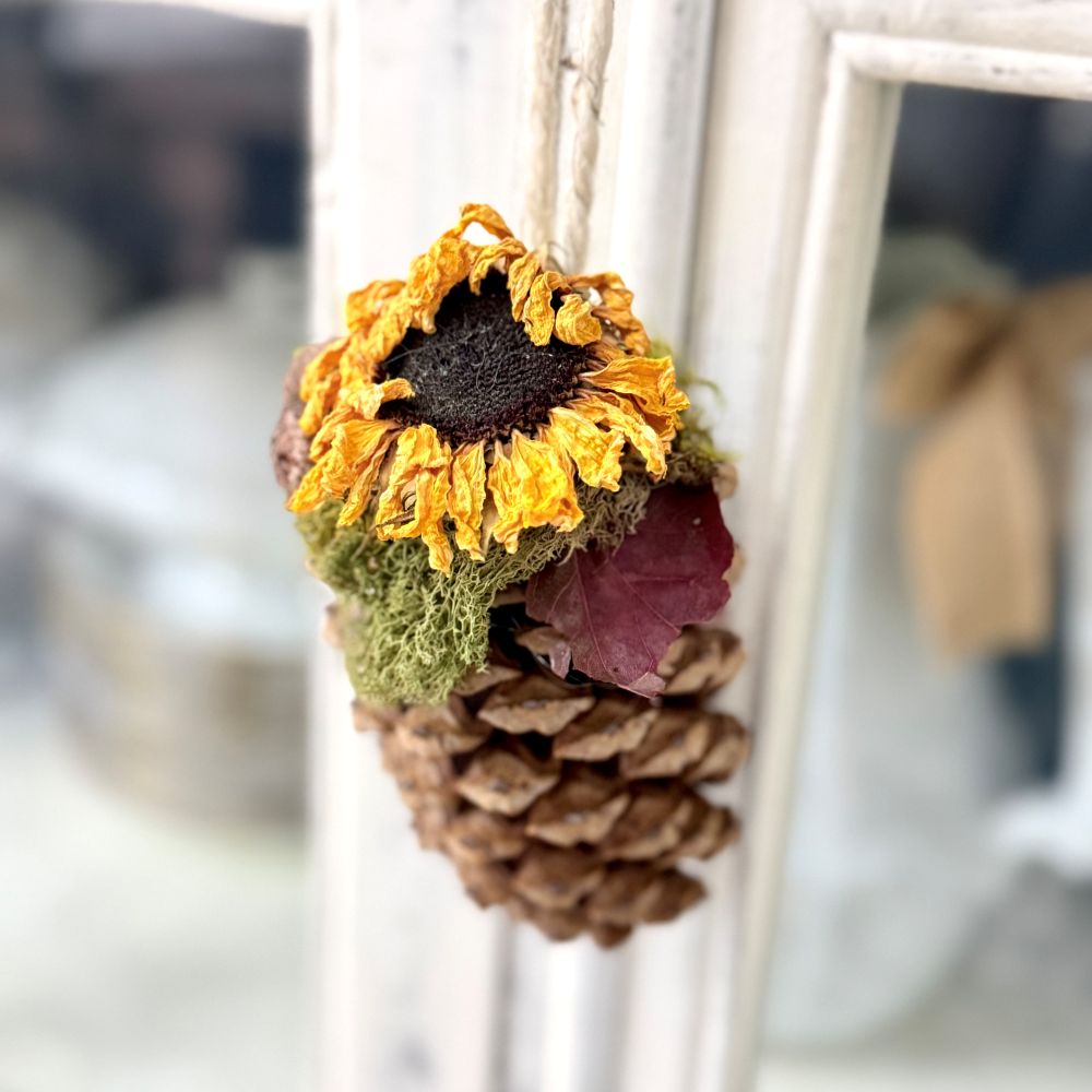 A hanging chair swag with a pinecone topped with moss, a dried sunflower, and a small red leaf.
