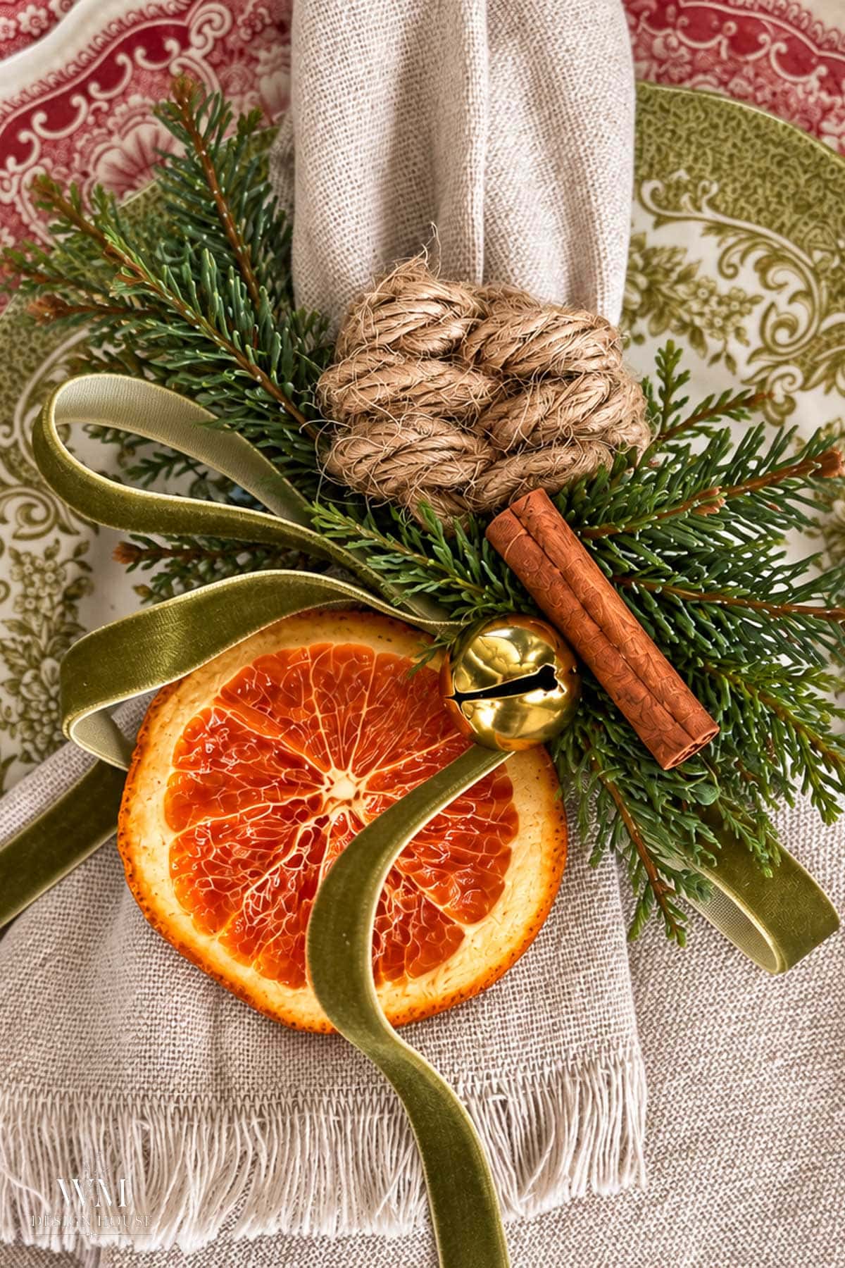 A linen napkin styled as a Christmas napkin ring, decorated with a dried orange slice, a cinnamon stick, evergreen sprigs, a gold jingle bell, and a leaf knotted rope twine.